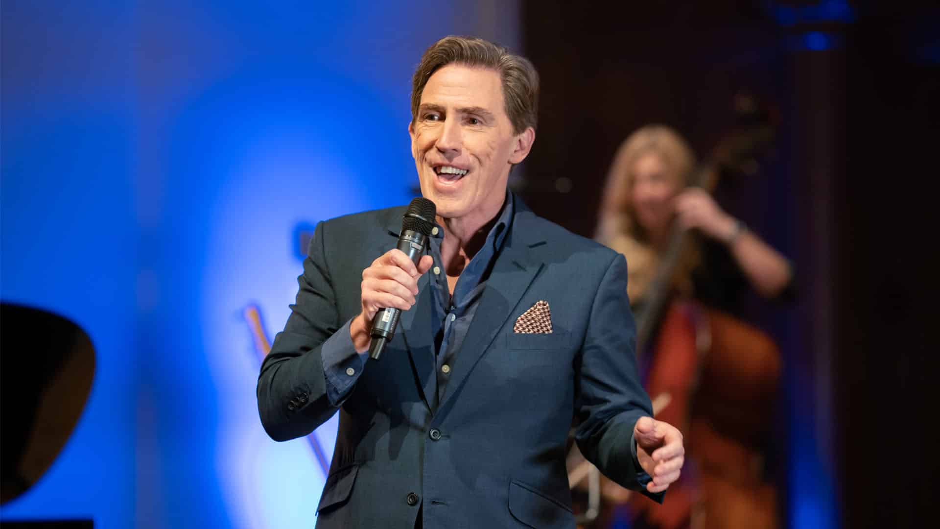 Production image of Rob Brydon on stage - smiling wide, microphone in hand