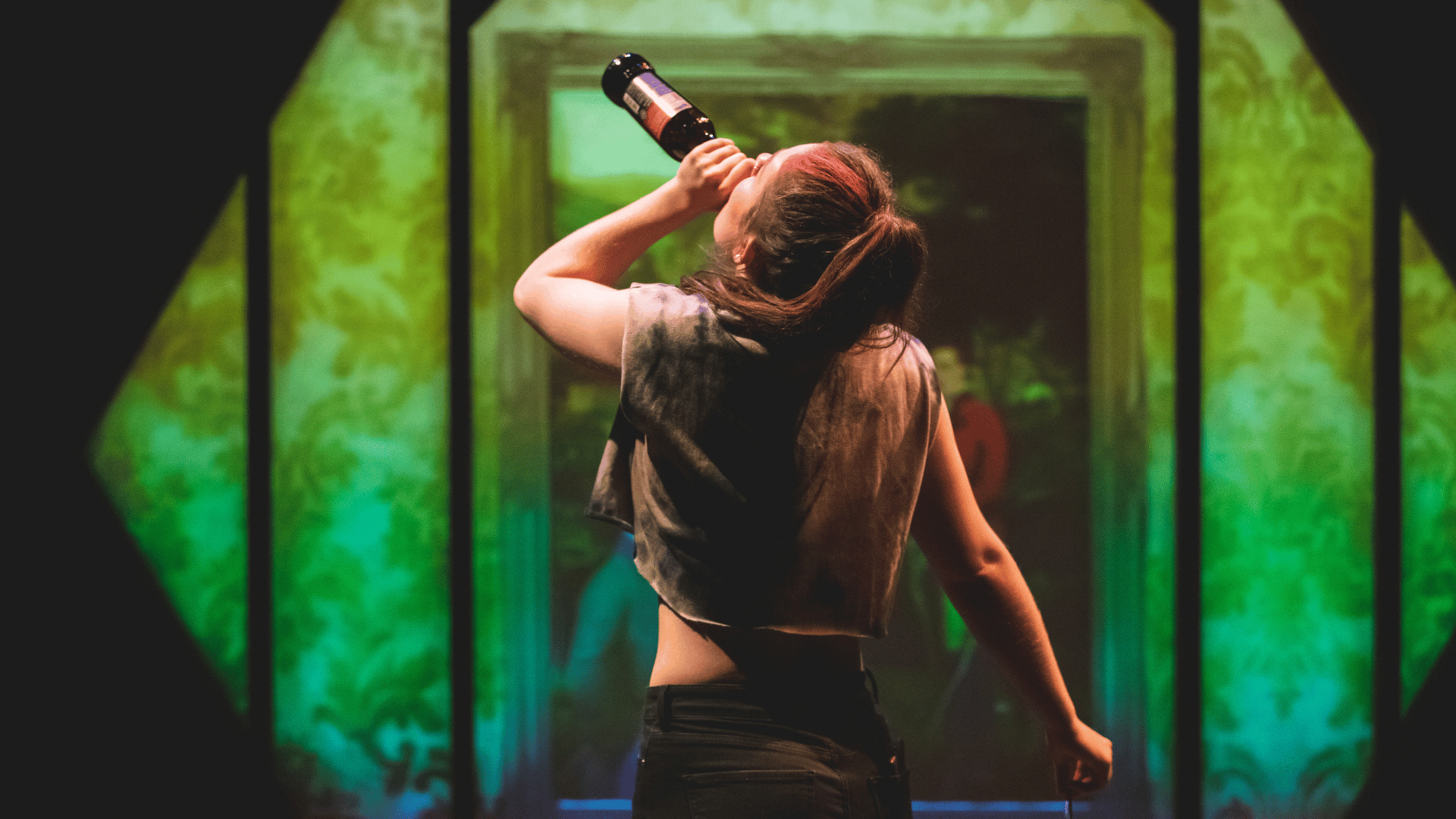 The Honey Man Production Photo: Misty (Amy Kennedy) dances and takes a swig from a bottle of beer, backlit by a colourful projection