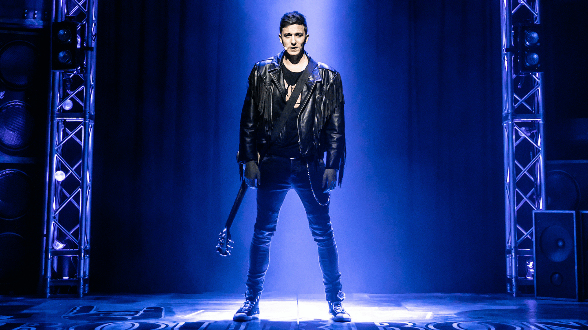 Rock of Ages production shot: the lead male actor stand alone on stage in a spotlight, carrying a guitar
