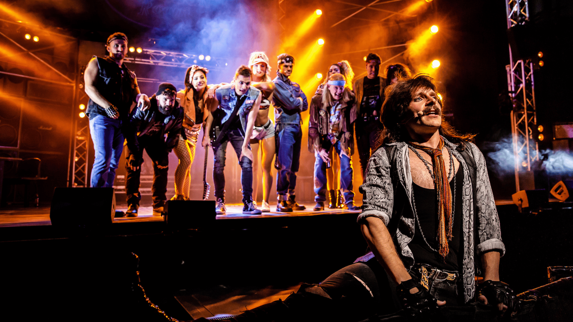 Rock of Ages production shot: a male actor sits on the edge of the stage, staring off into the audience, while the other cast members stand behind