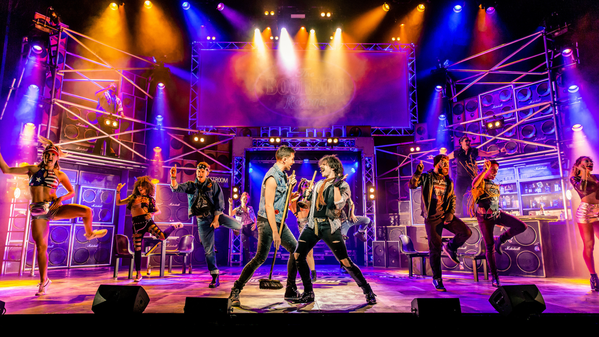 Rock of Ages 2018/19 tour production shot: Two male performers stand face to face, singing, while others dance in the background