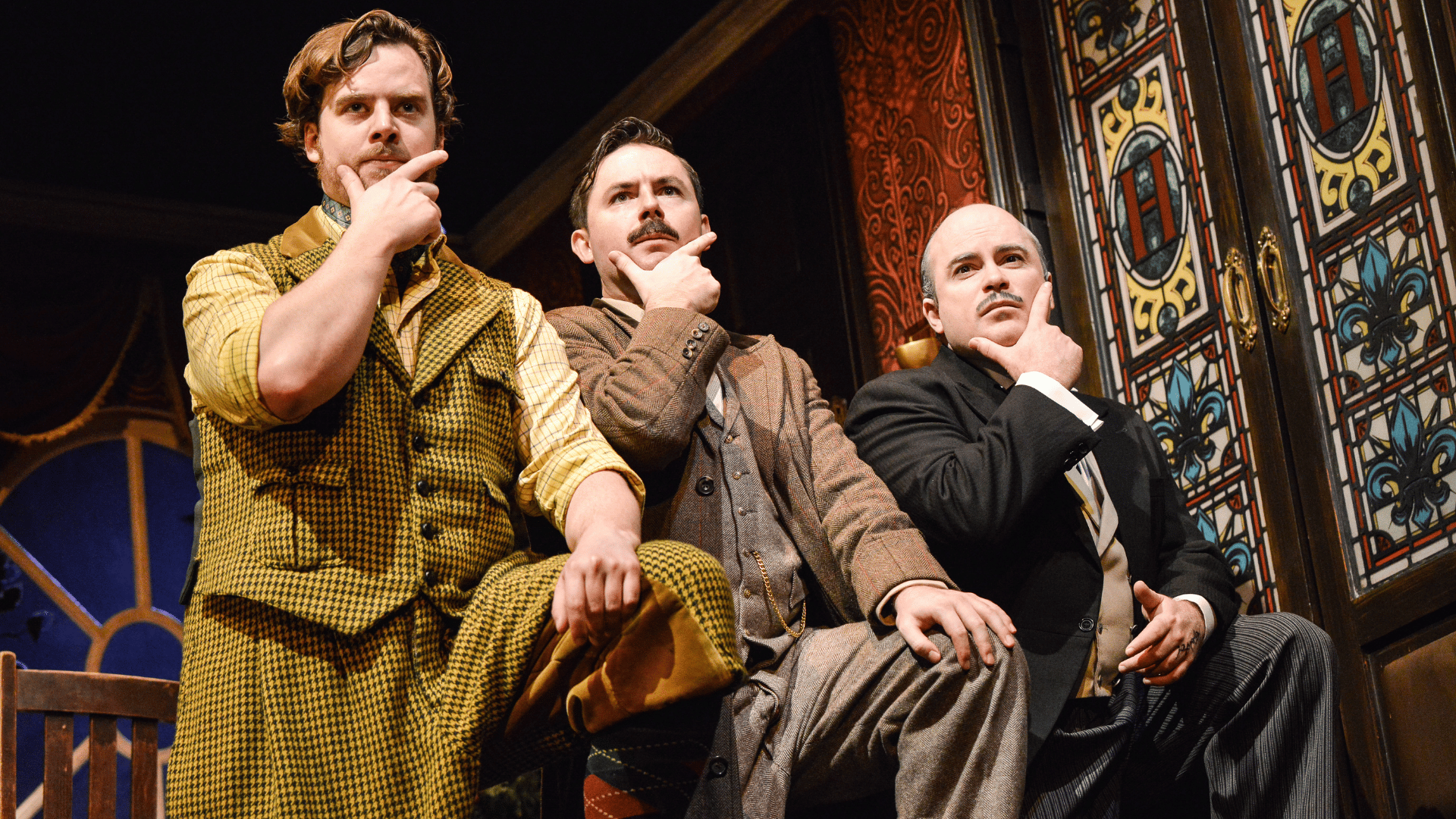 The Play That Goes Wrong Production Shot: Robert (Matthew Howell), Chris (James Watterson) and Dennis (Edward Howells) are standing next to each other all in the same position - one foot up on a rest, left hand resting on their left knee and their finger and thumb (right hand) stroking their chin.