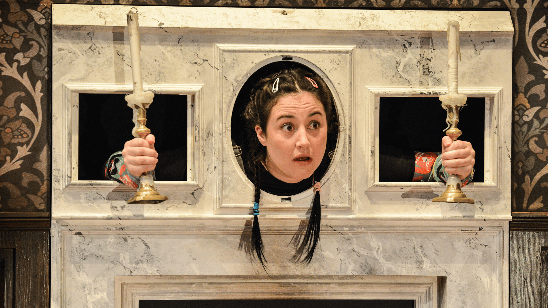 The Play That Goes Wrong Production Shot: A close up of Annie (Laura Kirman) who is stuck in a fireplace mantle piece, with her head through the middle hole and her arms through holes either side of her holding candlesticks.
