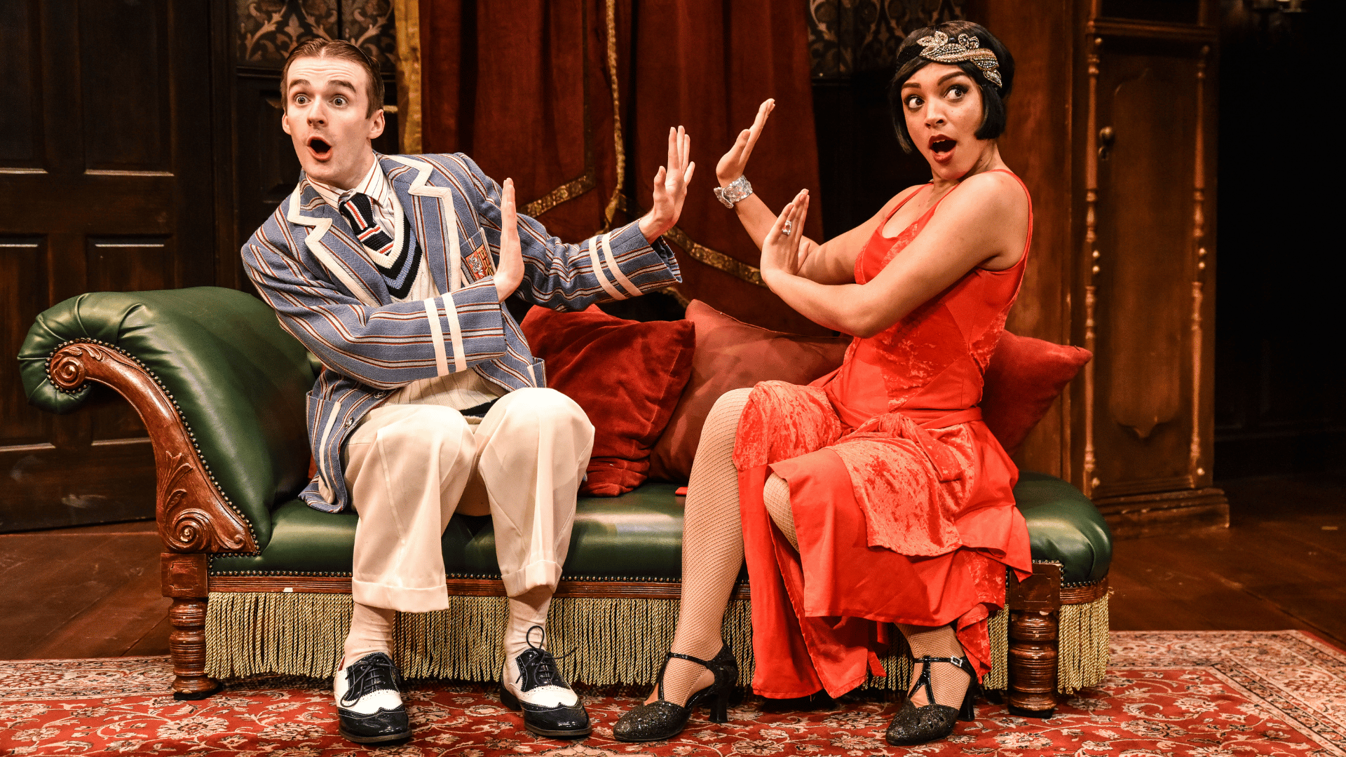 The Play That Goes Wrong Production Shot: Max (Tom Babbage) and Sandra (Laura White) are sat down on the chaise longue looking at the audience with a shocked face and their hands up as if they are pushing each other away.