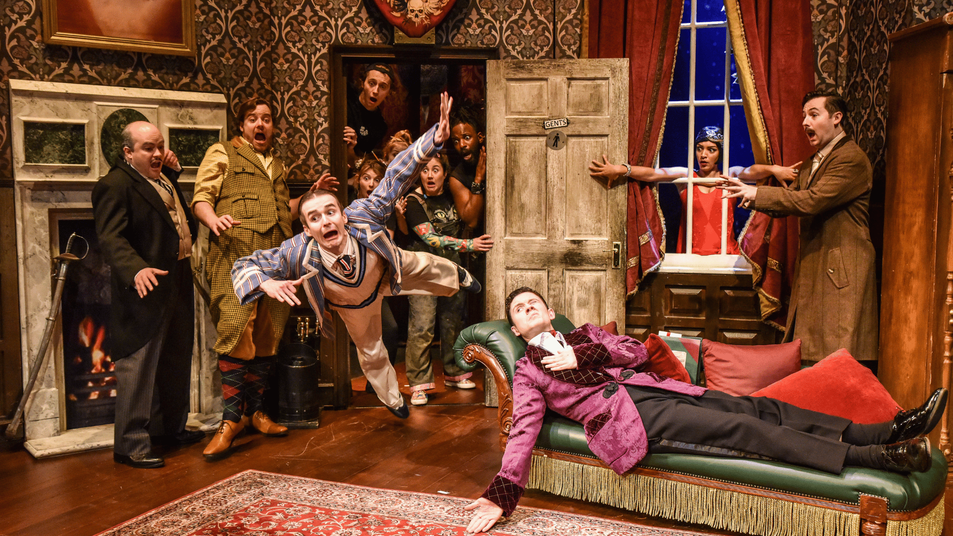 The Play That Goes Wrong Production Shot: Jonathan (Seán Carey) is laid down on a chaise longue with one eye open. Max (Tom Babbage) has burst into the scene falling through a door. Everyone else (9 cast members) are towards the back of the scene and looking scared as Max is falling.