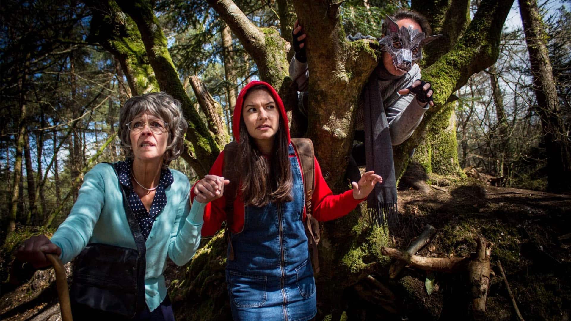 A modern Little Red Riding Hood and grandma are walking through the forest, concerned looks on their faces. A wolf is watching them from a tree right behind them, portrayed by an actor wearing grey clothing, a grey scarf, fingerless gloves and a very intricate and geometric wolf's mask