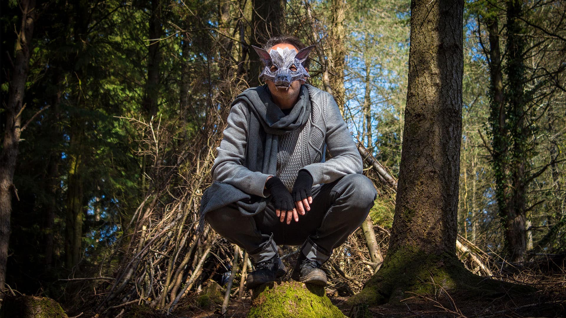 Little Red Riding Hood's Wolf is sitting crouched on a boulder in the woods,portrayed by an actor wearing grey clothing, a grey scarf, fingerless gloves and a very intricate and geometric wolf's mask