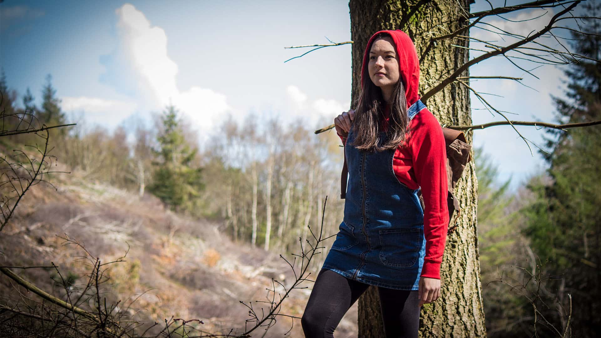 A modern-day Little Red Riding Hood is walking in the woods, wearing a blue jean overall dress with a red hoodie underneath, long brown hair peaking from under the hood. She's looking really determined.