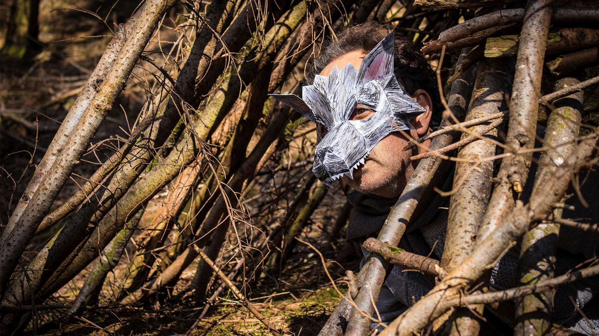 The Big Bad Wolf hiding between branches, portrayed by an actor wearing grey clothing, a grey scarf, fingerless gloves and a very intricate and geometric wolf's mask