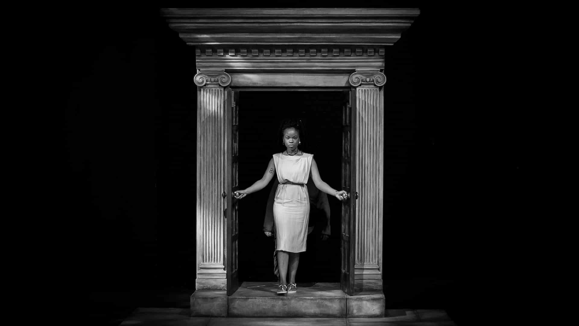 Antigone Rehearsals - Rachel Nwokoro as Antigone, defiantly walking through an ancient looking pillared doorway, wearing a tunic dress
