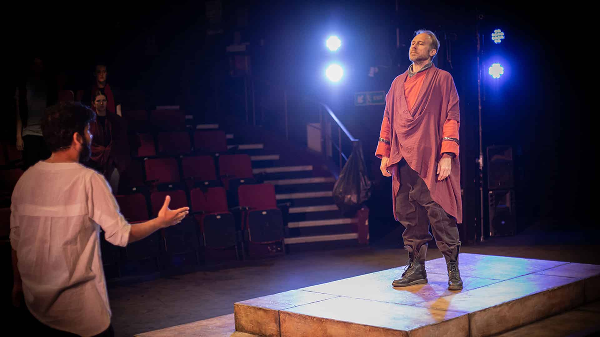 Antigone Rehearsals - David Annen as Kreon on a small platform on stage, wearing a modern toga style costume and combat boots, being directed by Associate Director Yusuf Niazi