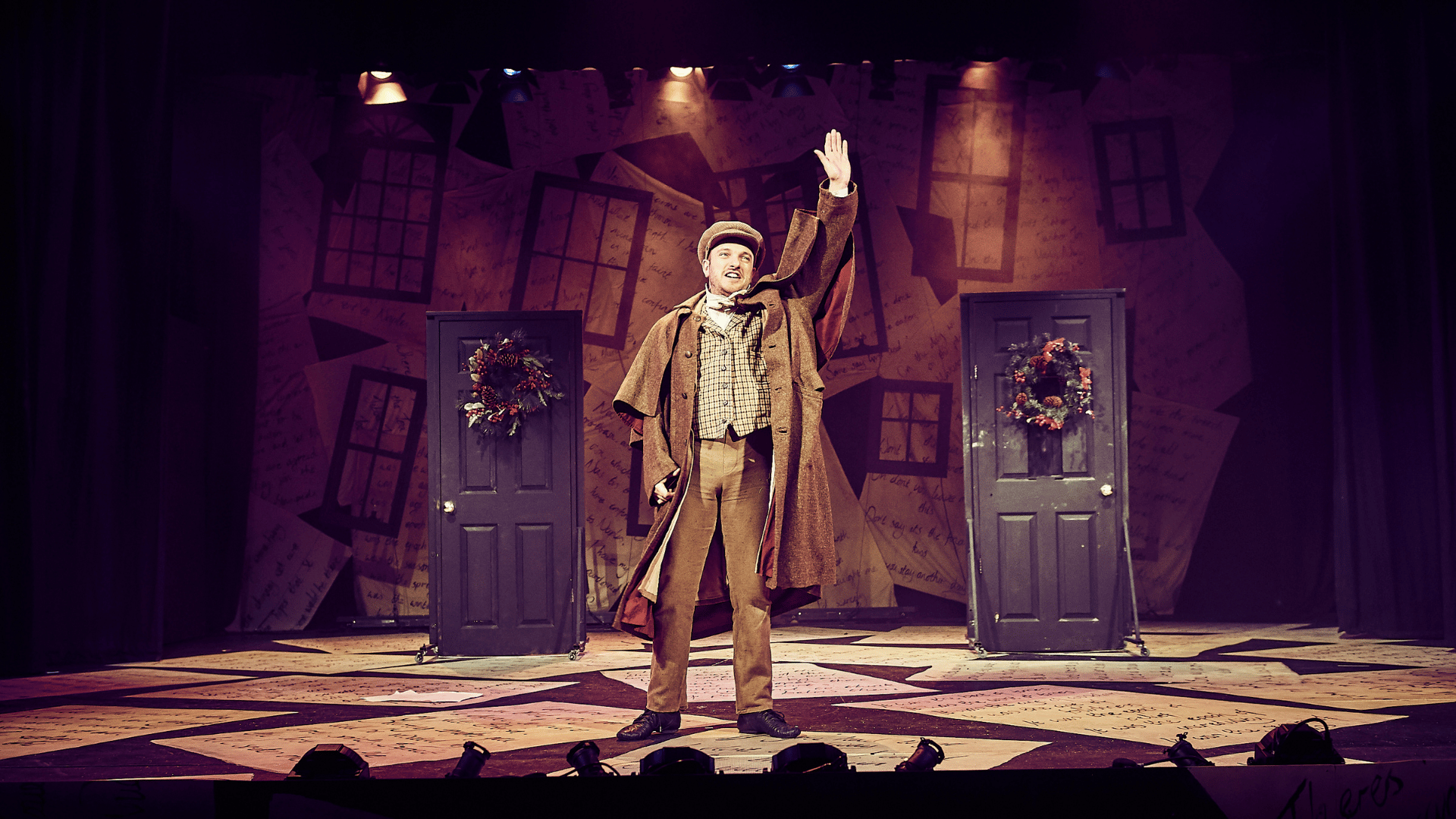 A Christmas Carol production shot: a character is stood in the centre waving at the audience. In the background, there are 2 doors decorated with Christmas wreaths