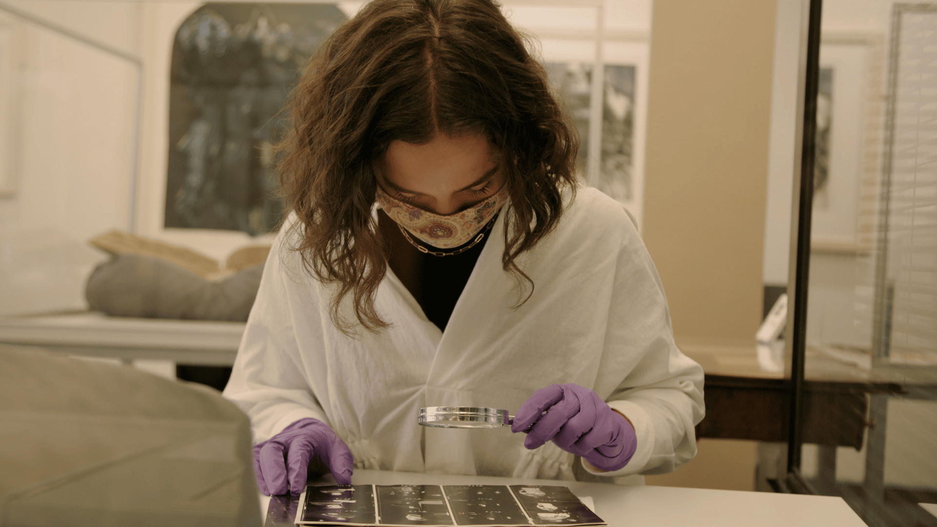 A female intern dressed in a lab coat, gloves and goggles uses a magnifying glass to look at photo negatives in the archive