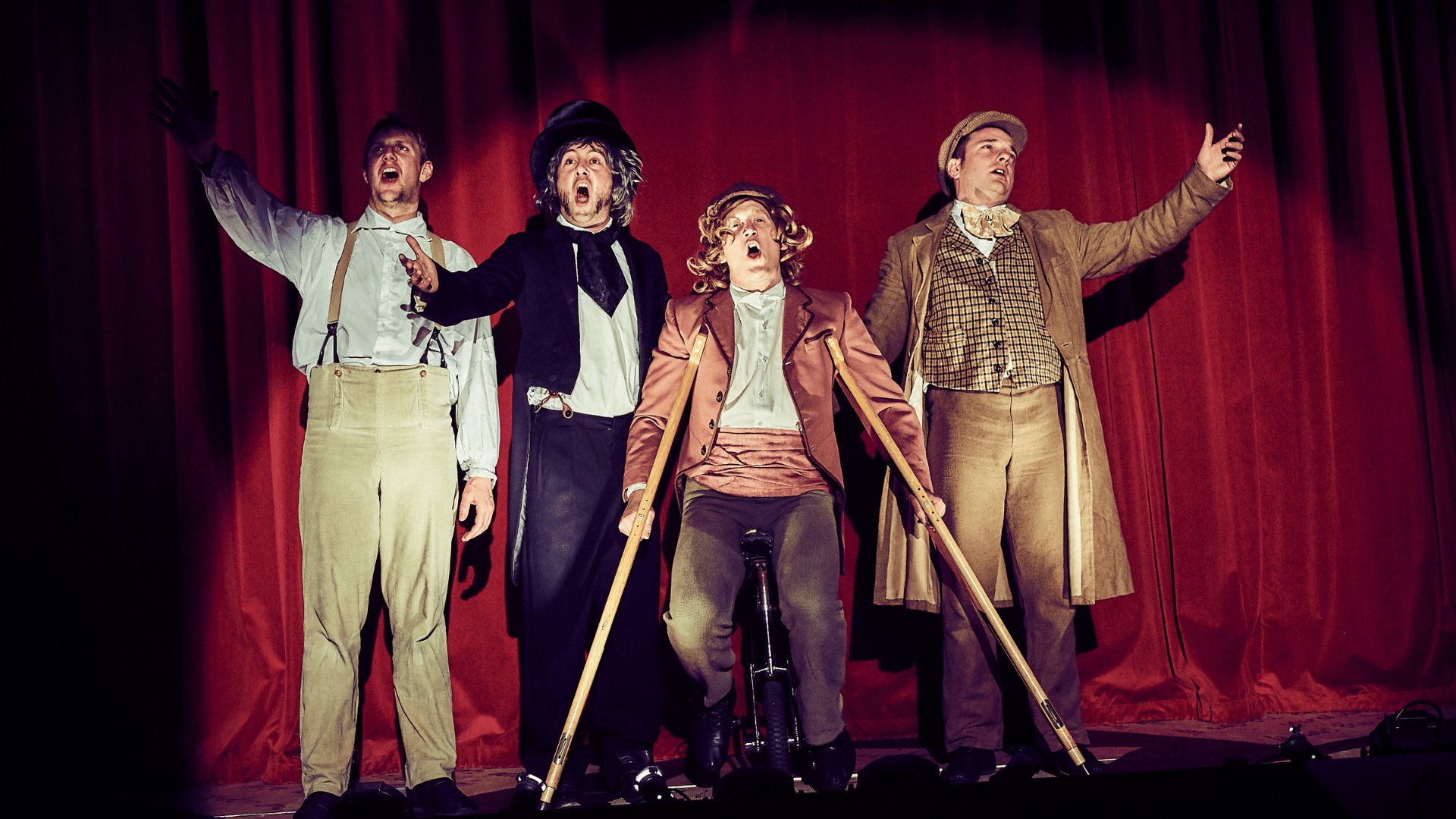 A Christmas Carol production shot: 4 character stood in front of the curtain singing. 1 character is on crutches.