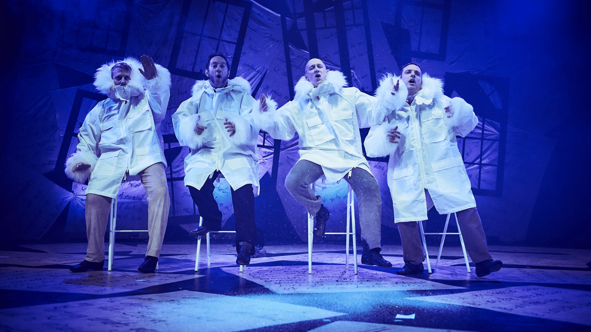 A Christmas Carol production shot: 4 characters sat on stool, wearing large white winter coats