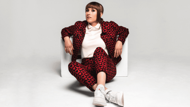 MAISIE ADAM - BUZZED artwork. Maisie Adam sitting on the floor leaning back against a box. She wears a matching black and red jacket and trousers. Her hair is shoulder length at the front and shaved at the back.