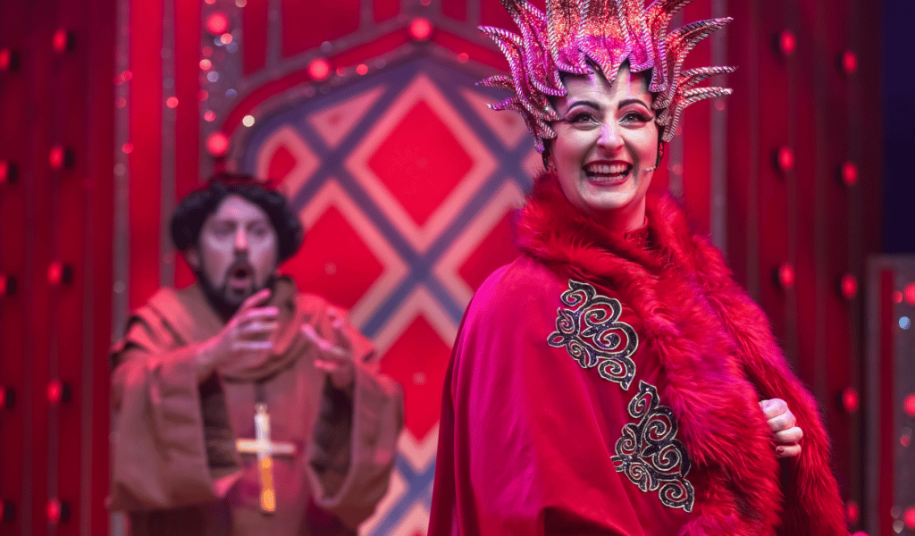 In the foreground, Jodie Micciché as Morgana cackling wickedly while wearing a red gown with fur trim and a sparkly, pointy red headdress. Out of focus in the background, Nick Bunt as Merlin in disguise as Friar Tuck, wearing a monk's brown robes and a crucifix, looking towards Morgana with an aghast expression on his face.