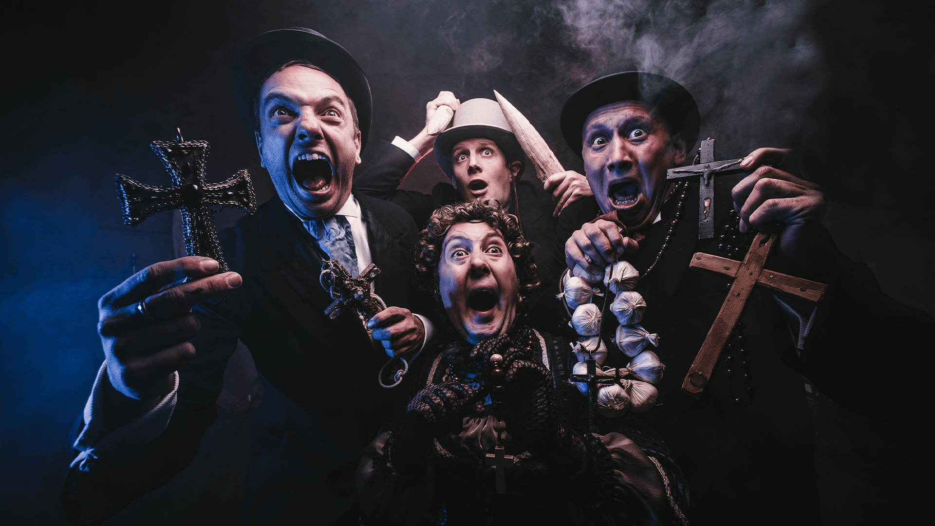 Black, smoke-filled backgrond. Four members of Le Navet Bete scream in horror at the camera. The performers on the left-hand side of the image is wearing dark clothing, a top hat and holding two crosses. The performer on the right-hand side is wearing dark clothing and a top hold and is holding garlic and two crosses. The performer at the bottom of the image is holding roasary beads and wearing velvet gloves. The performer at the top of the image is waving wooden stakes above their head.