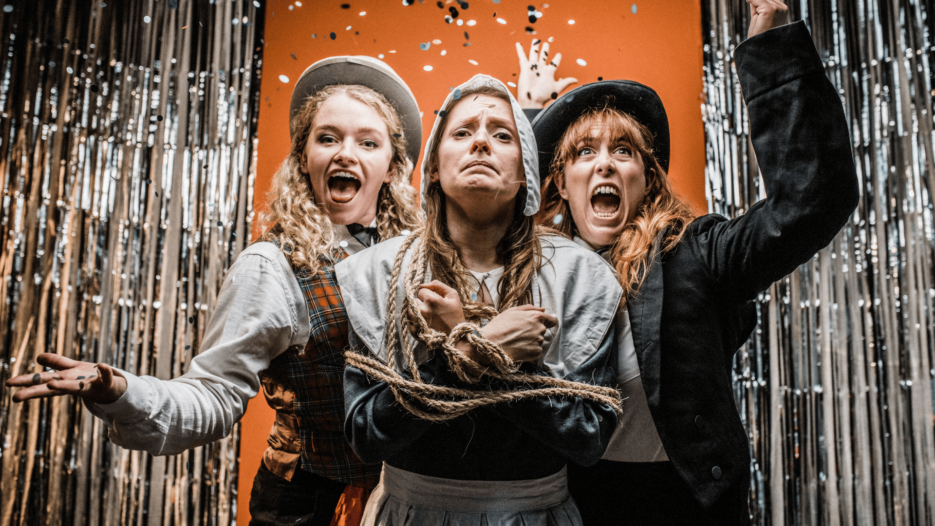 Hags: A Magical Extravaganza artwork. Three young women (left and right, dressed as stage magicians; centre, dressed as a medieval peasant and bound with a rope) stand between two tinsel curtains against an orange background. The two women dressed as magicians have shocked expressions and the woman dressed as a peasant looks worried.