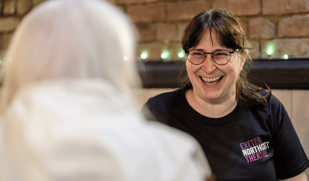A woman, wearing an Exeter Northcott Theatre-branded black t-shirt, laughs with a visitor.