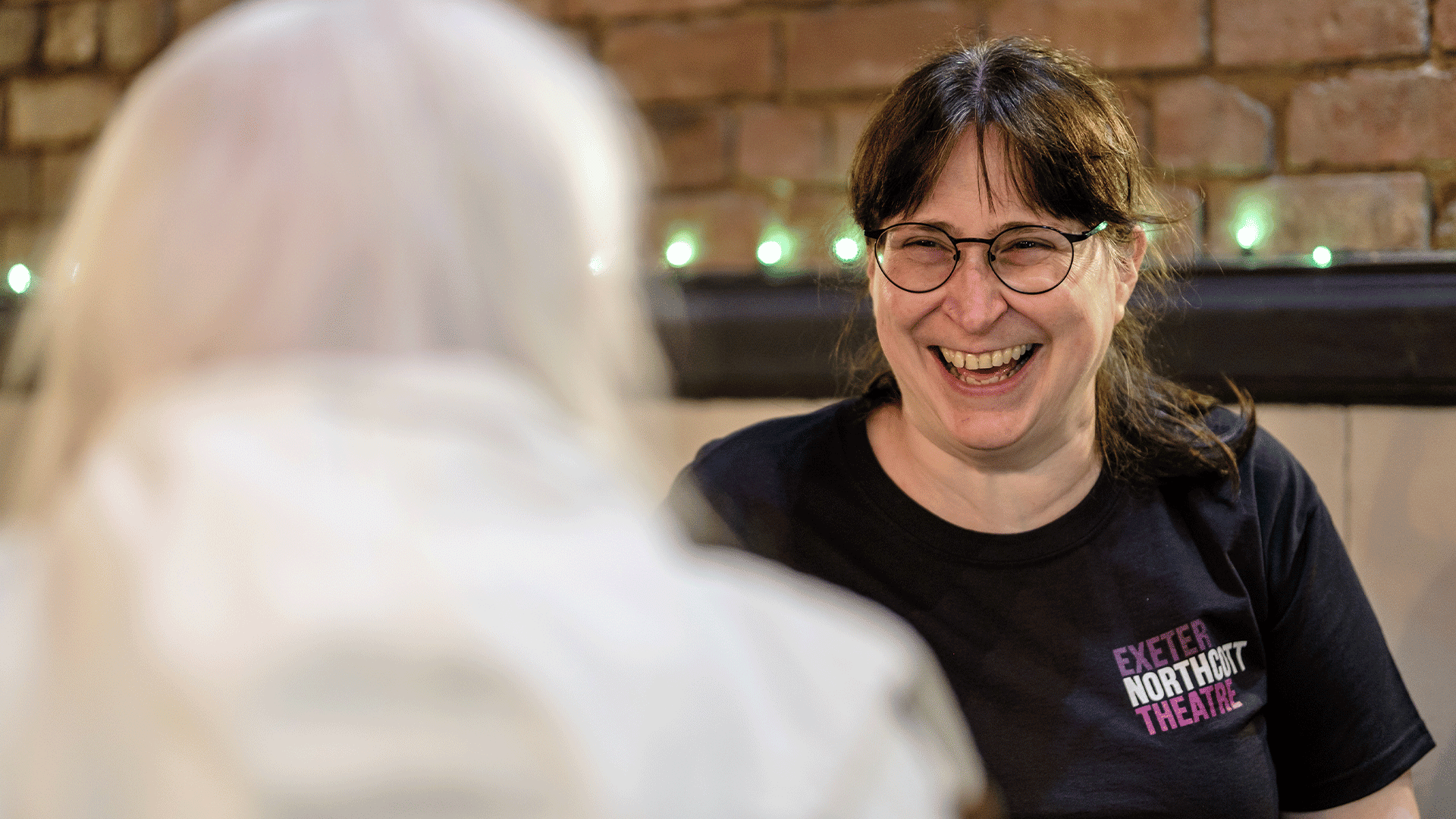 A woman, wearing an Exeter Northcott Theatre-branded black t-shirt, laughs with a visitor.