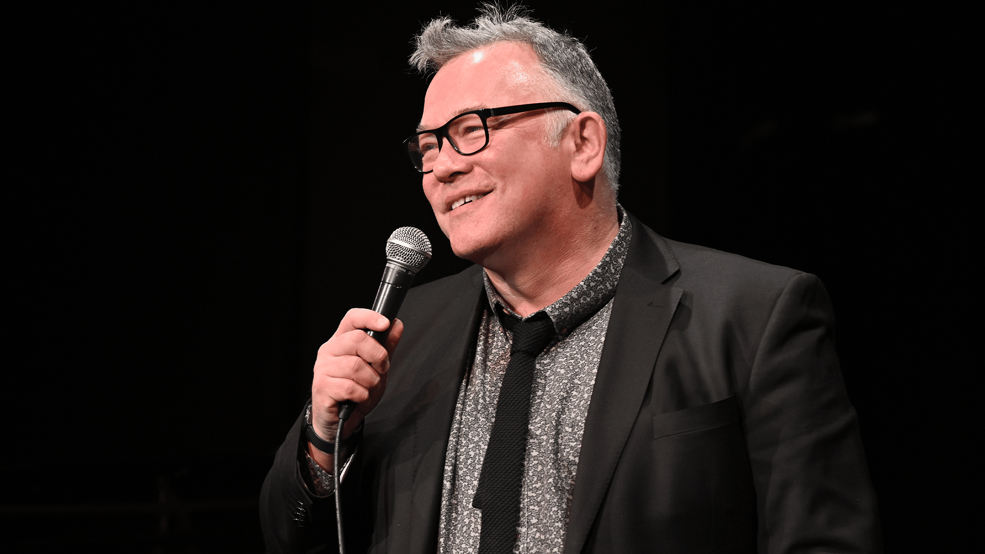 Stewart Lee wearing a black suit, holding a microphone and smiling