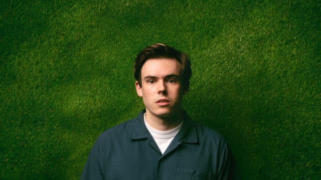 Rhys James stood facing the camera in a blue shirt in front of a dark green background
