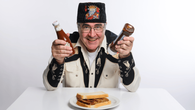 Image of Danny Baker sitting at a table. A white bread sandwich with sausages in front of him. He's holding brown sauce in one hand, red sauce in the other