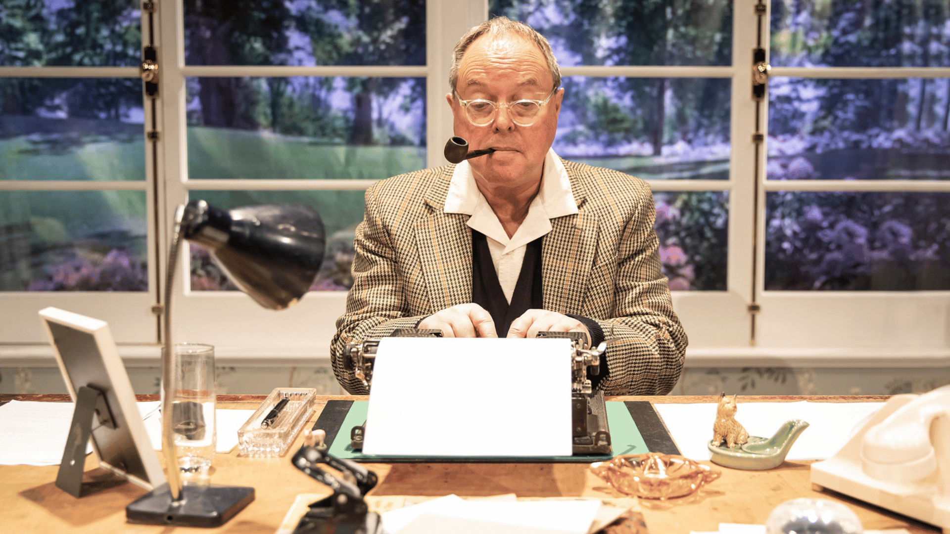 Robert Daws as PG Wodehouse sat at his desk typing into a typewriter. He has a smoking pipe in his mouth and he is wearing a tweed jacket with a white shirt and black waistcoat underneath. His desk contains a lamp, telephone, picture frame and various trinkets. Behind him is a window with a nice view of trees, flowers and grounds.
