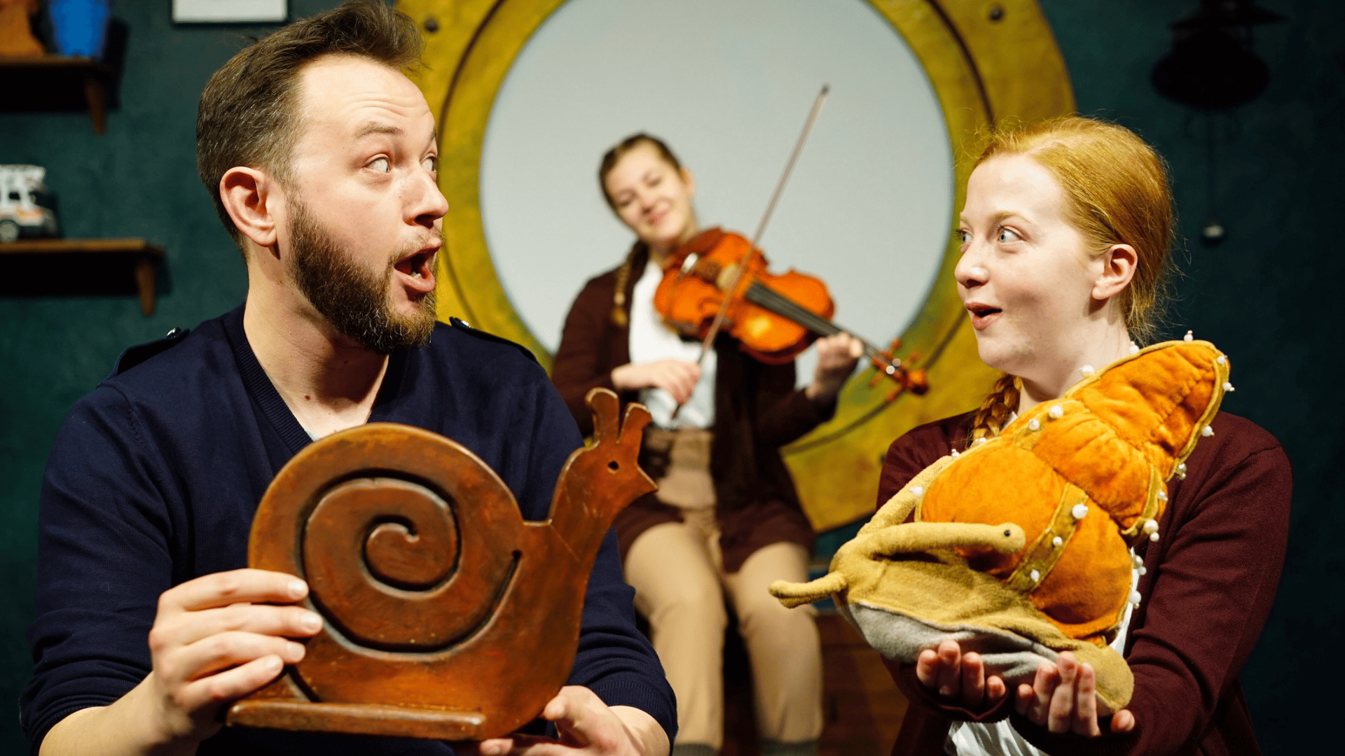 A man holding a wooden snail faces a girl holding a soft toy sea snail. In the background a female violinist plays in front of a large porthole.