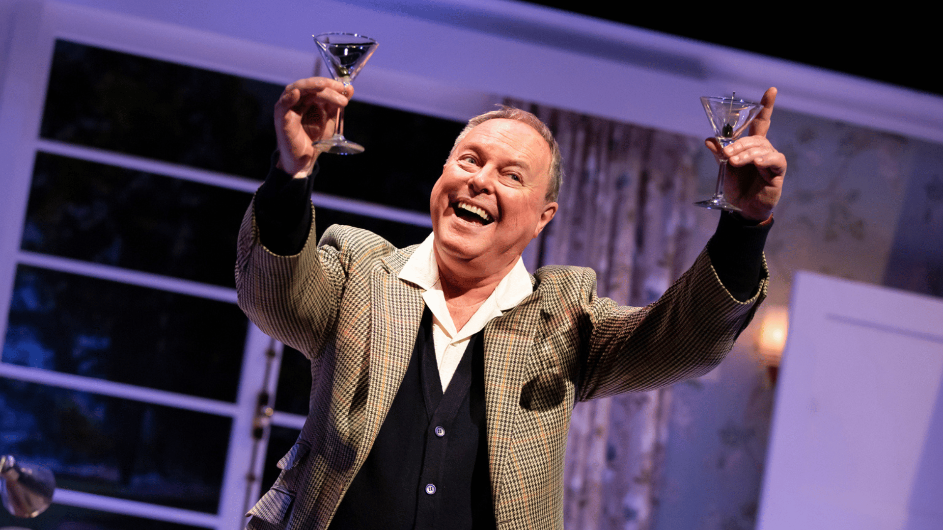 Robert Daws as PG Wodehouse holding two martini glasses on the air with a wide open-mouthed smile on his face. He is wearing a tweed jacket with a white shirt and black waistcoat underneath.