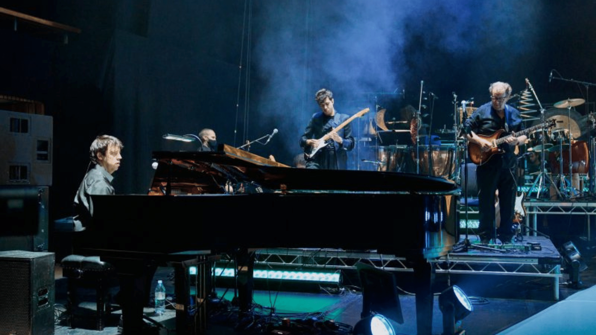 A man playing at a black grand piano. In the background, two men play electric guitars.