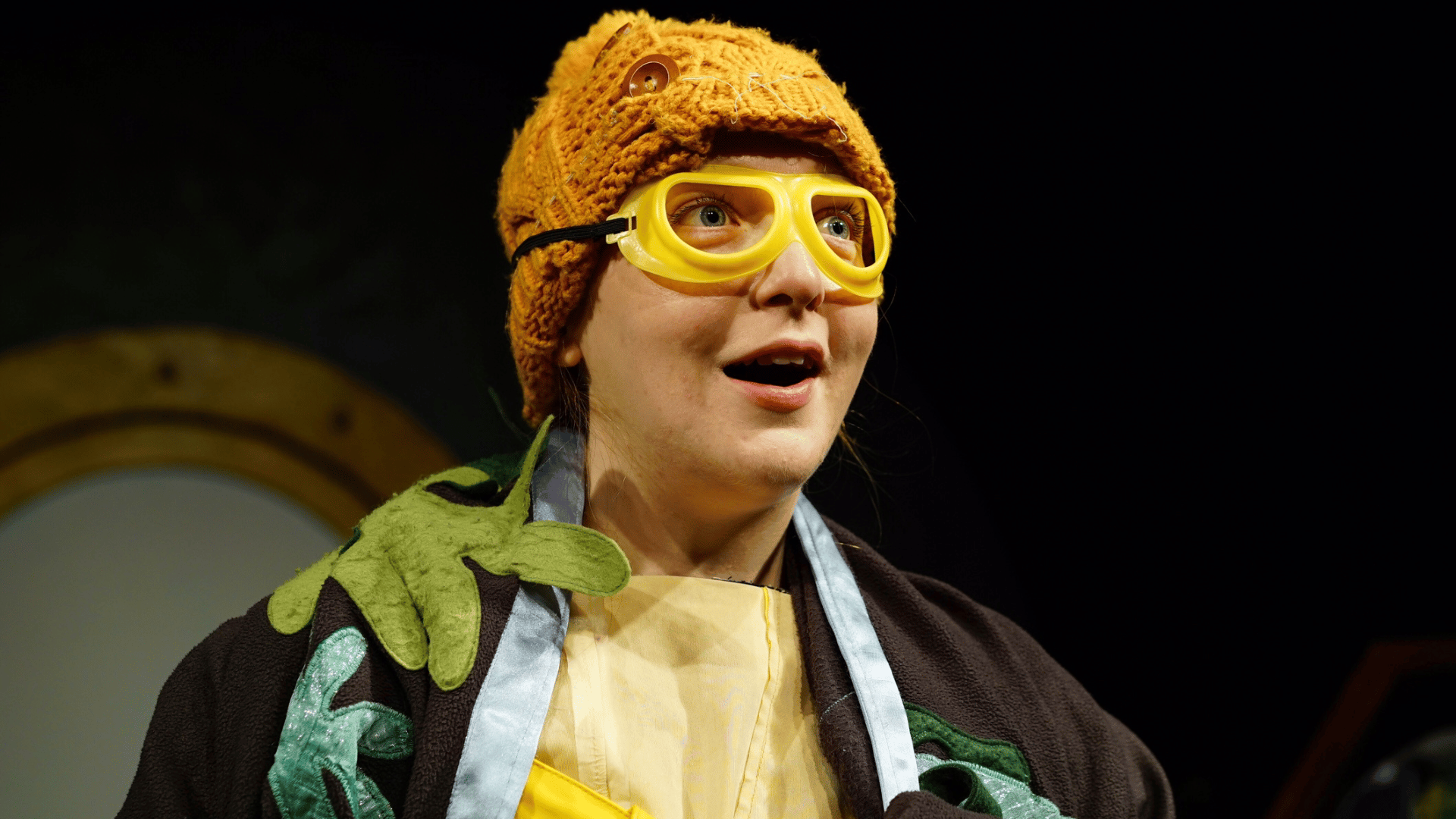 A person in a mustard-yellow beanie hat with yellow goggles on, staring ahead with a look of awe on their face.
