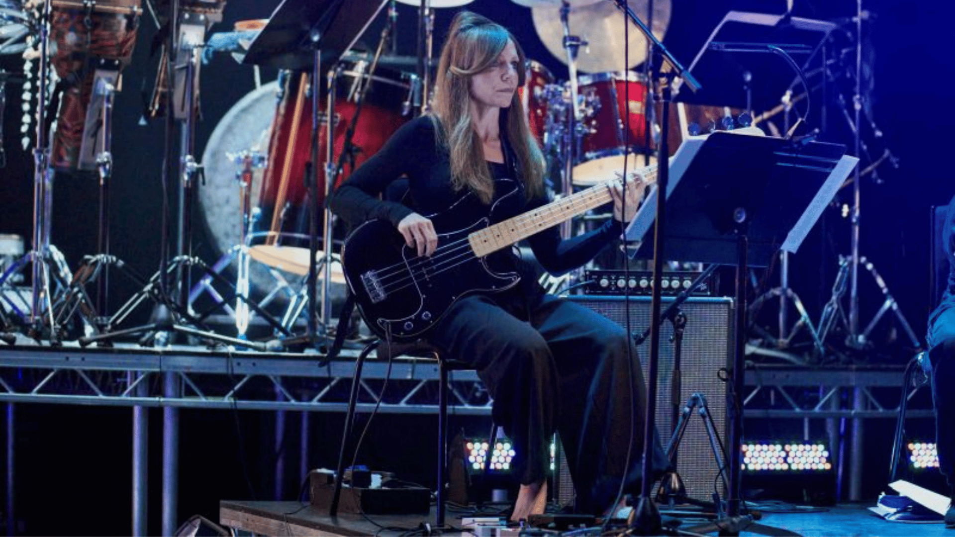 A woman in a black outfit, sat in front of a drum kit playing a bass guitar.