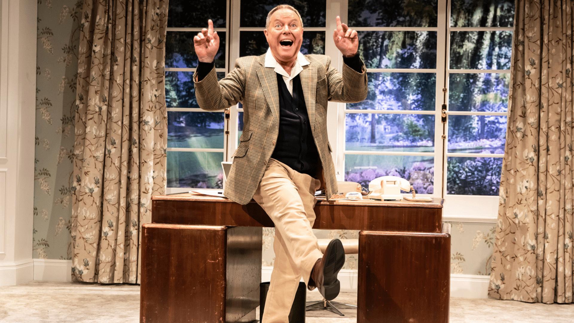 Robert Daws as PG Wodehouse pointing his index fingers in the air and lifting his right leg out in a dancing pose. His mouth is wide open with a happy expression on his face. He is in front of his desk and a window with a nice view of trees, flowers and grounds.