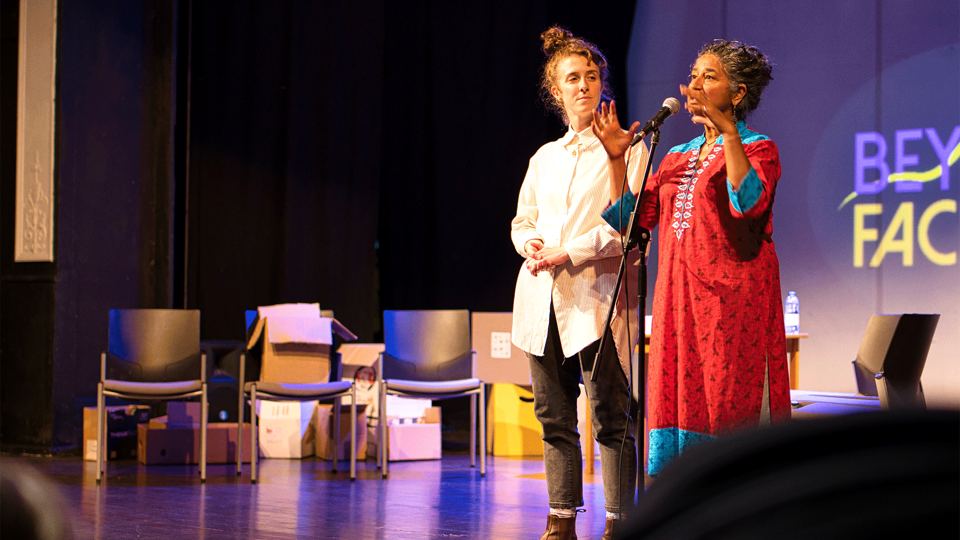Northcott Community Producer Nicole Redfern and Devon Diversity Consultant's Sandyha Dave address an audience from the Barnfield auditorium stage. Behind them, a series of chairs and cardboard boxes, and the Beyond Face theatre company logo. Text reads: 'Beyond Face'.