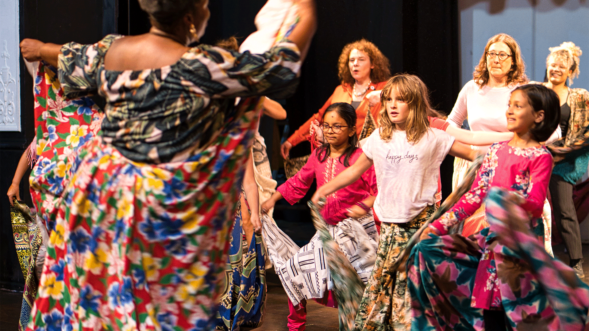 A woman holding colourful fabric behind her raises her arms in front of a group of children and adults, who are also holding fabric and raising their arms.