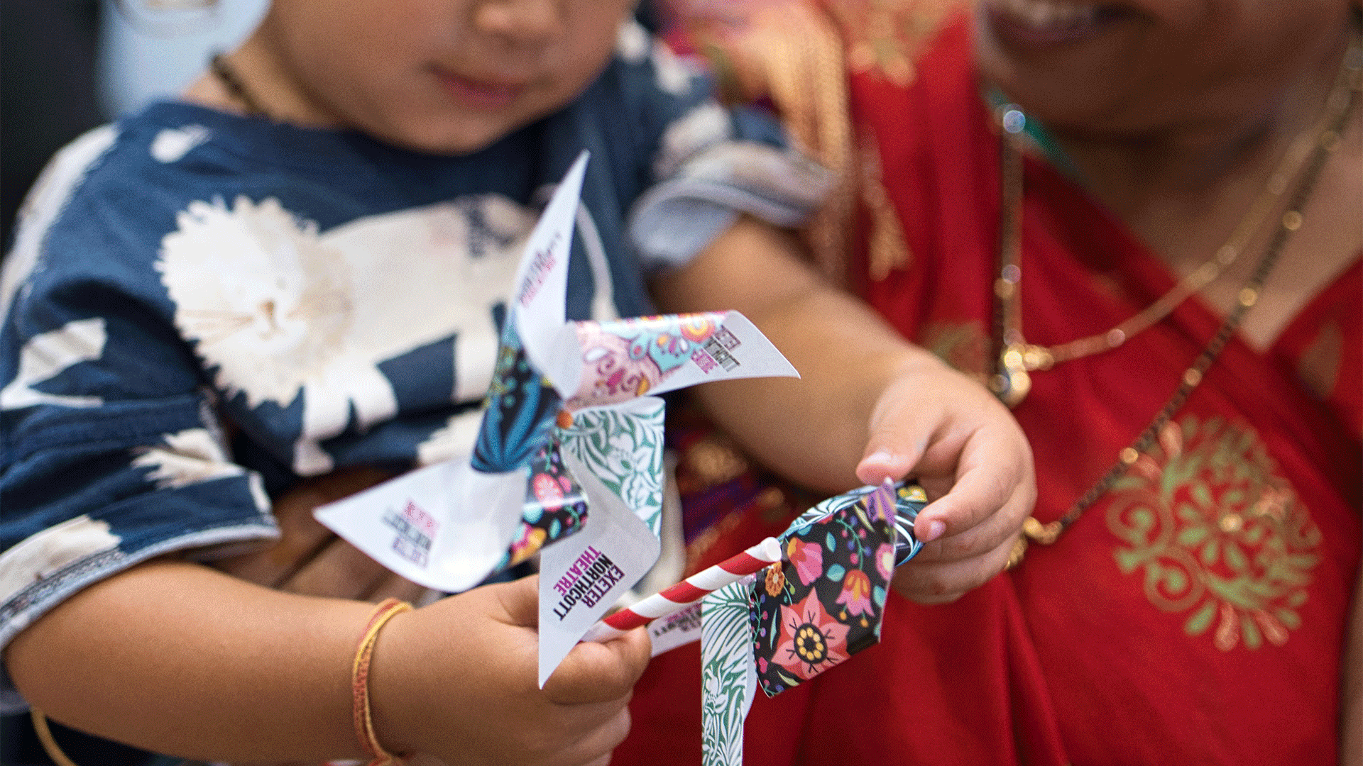 A young girl, held in arm by a woman in a red and gold dress, holds up a paper windmill featuring patterns from all of the communities involved in The Story of Us festival as well as the Exeter Northcott Theatre logo.