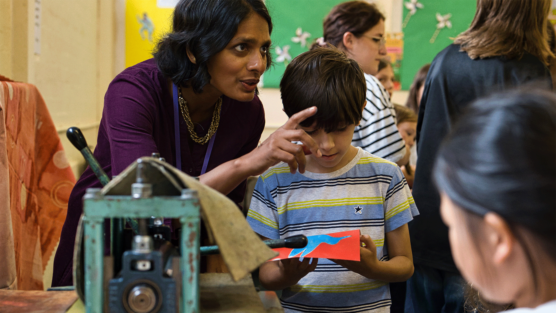 A woman points to her left while talking to a child, who is looking down at a fabric print held in their hands. In front of them, a printing press.