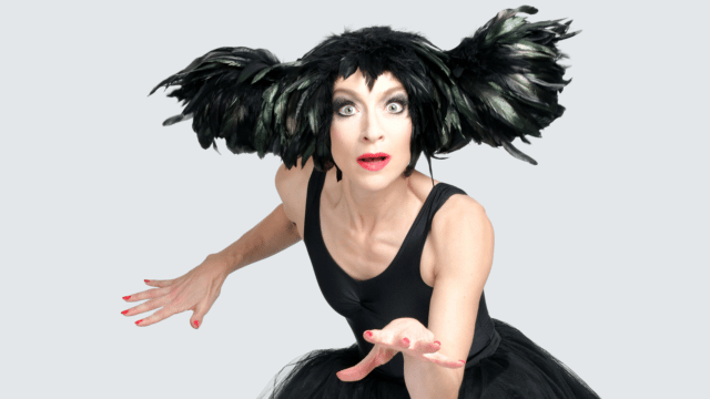 An Evening Without Kate Bush - photo by Steve Ullathorne. Sarah-Louise Young (dressed in a black tutu and feathered black headdress) is caught jumping in mid-air, her legs tucked up underneath her and her hand outstretched towards the camera. She looks into the camera with a look of surprise on her face.