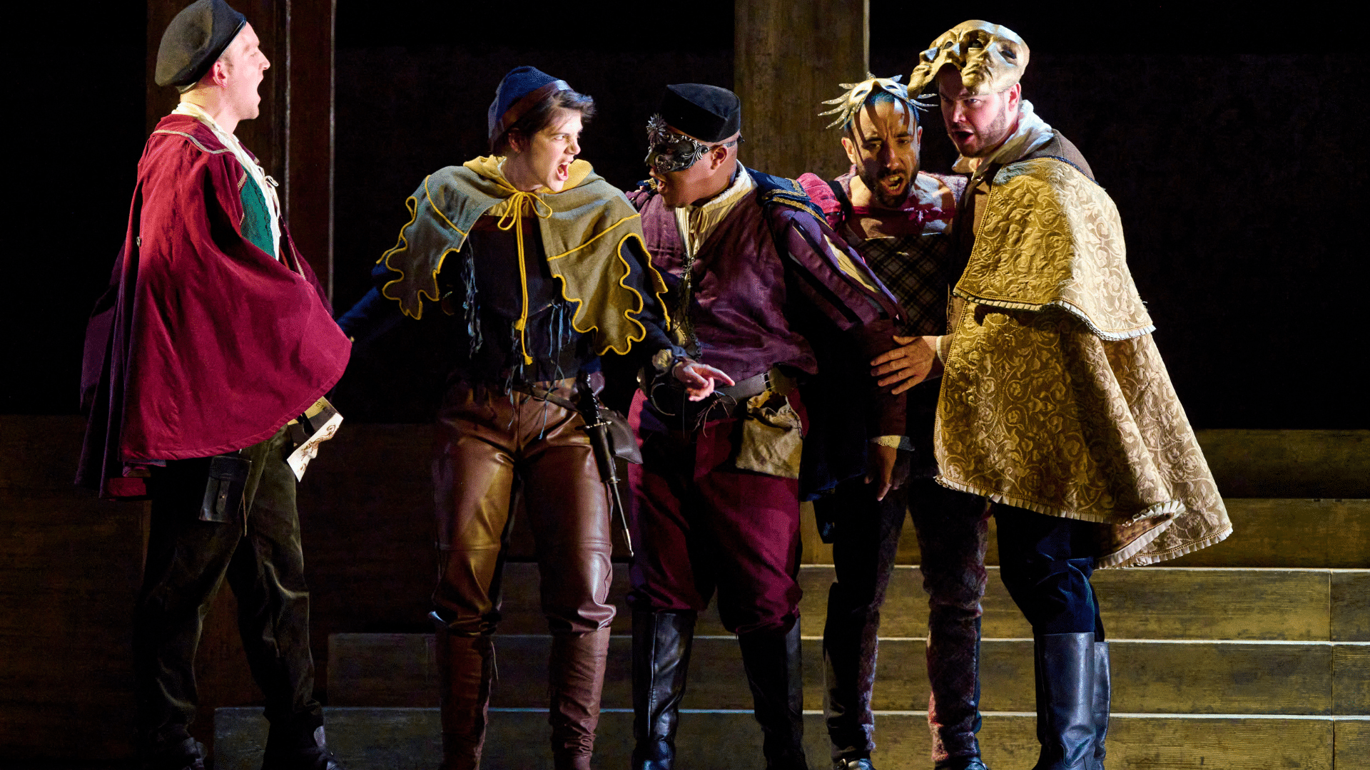 ETO: Lucrezia Borgia production shot - A group of five men wearing colourful outfits stood closely together in a line. They are all singing and they have serious expressions on their faces. The man in the middle wears an ornate mask across his eyes and the two men on the right have masks resting on their heads. Behind them are some wooden steps.