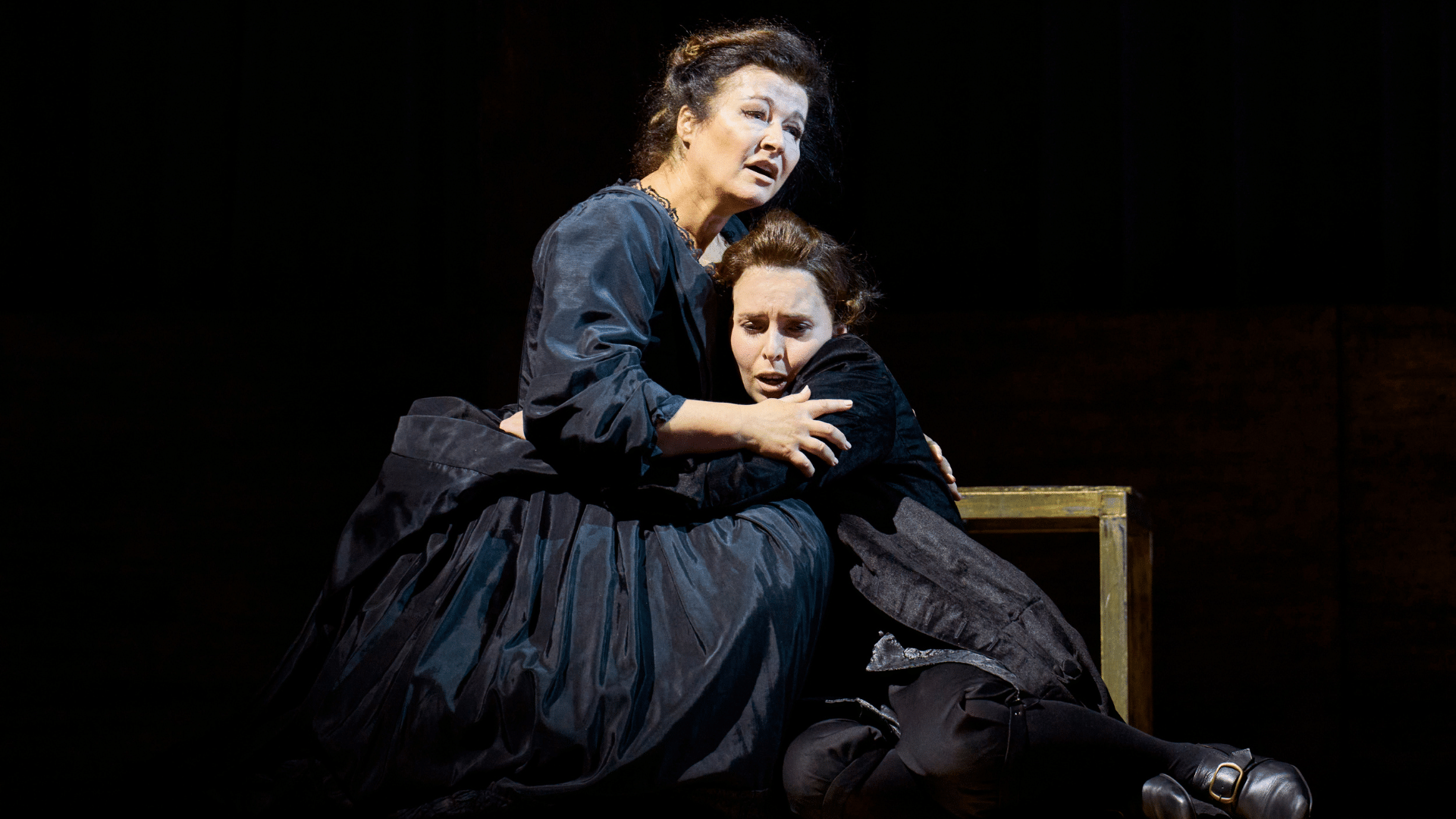 ETO: Giulio Cesare production shot - Two women in black dresses embrace while kneeling on the ground. They look sorrowful. A small wooden table is just behind them and the background is completely black.