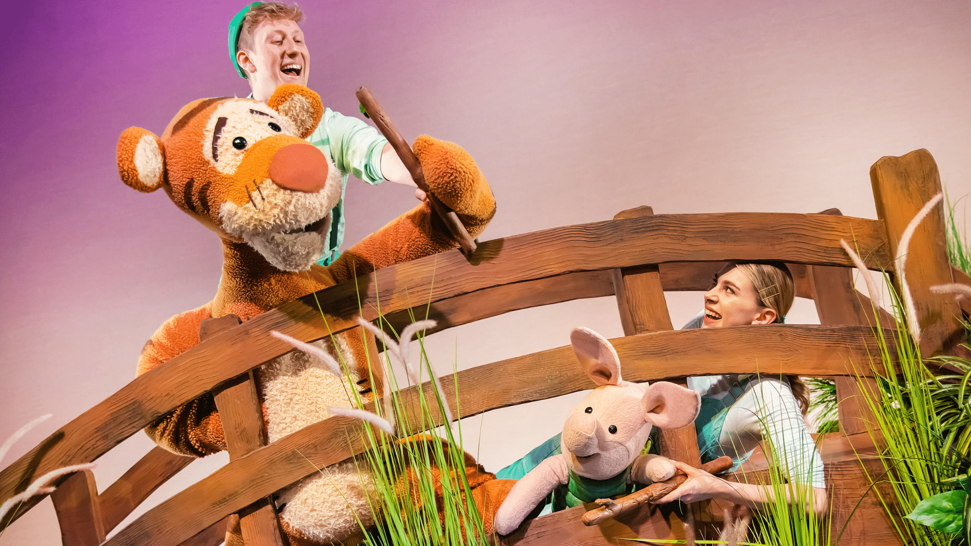 Disney's Winnie the Pooh production photo - Tigger and Piglet puppets in a wooden bridge above a blue stream with green reeds sprouting from the water. Tigger leans over the top of the bridge with a wooden stick in their hand. Piglet peeks out from the bottom of the bridge, also holding a stick. Two actors operate the puppets.