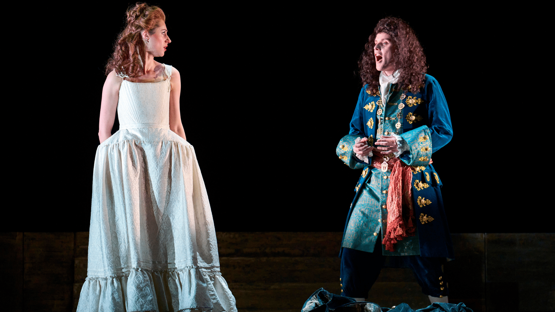ETO: Giulio Cesare production shot - On the left, a woman with red hair and red lipstick is wearing a pretty white dress, with her hands hidden behind the middle of the dress. She faces to the right where a man with long, dark, curly hair sings while facing her. He wears a very grand blue coat decorated with ornate, golden leaves and a long red scarf wrapped around his waist. The background is completely black.