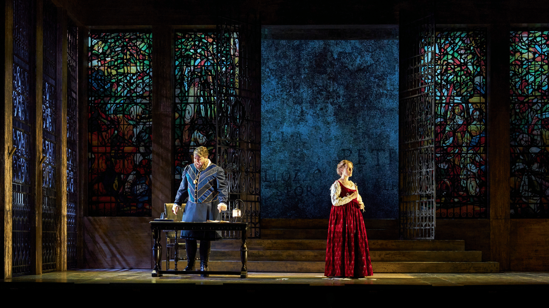 ETO: Lucrezia Borgia production shot - On the left, a man wearing blue and black striped tunics stands behind a desk, while yelling as he faces downwards. On the right, a woman in a red floor-length dress with a white long-sleeved under-blouse faces away from the man towards the right. Behind them on both sides are large, colourful stained glass windows and in the middle, steps leading to an opening flanked with black metal gates. Through the opening is a blue textured wall with some roman lettering.
