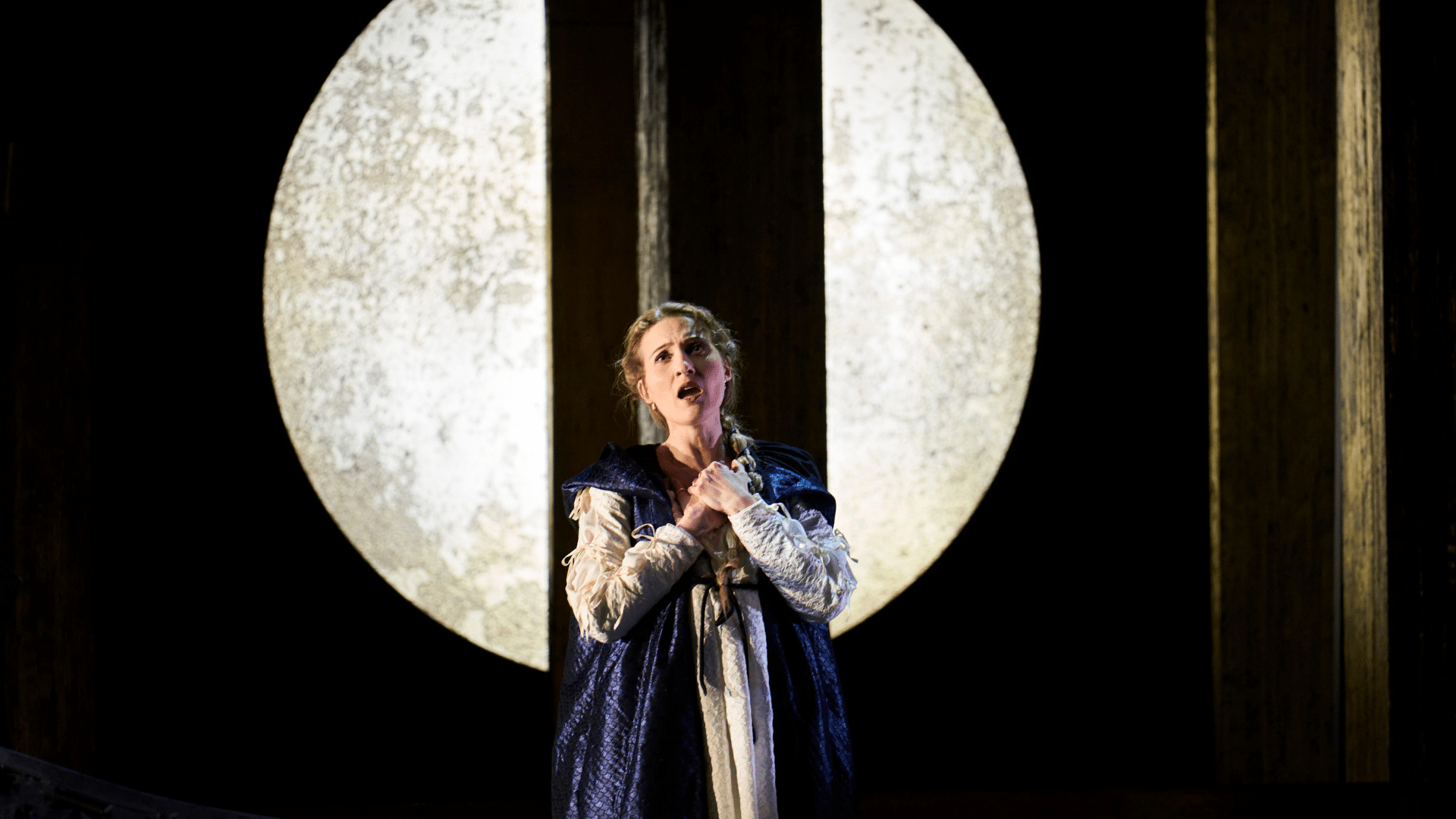 ETO: Lucrezia Borgia production shot - A woman wearing a blue robe with a white long-sleeved under shirt, faces forwards and holds her hands together across her chest while singing. Behind her is a white glowing moon-like orb, split in two to form an opening.