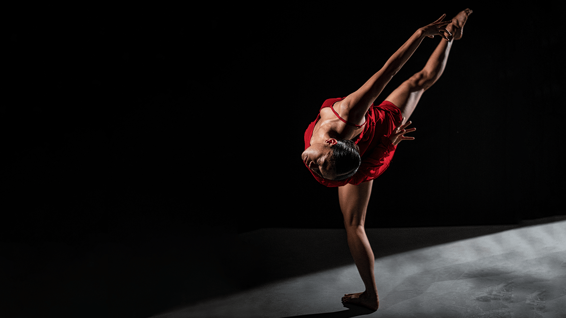Hot House promotional image (photo by Jon Bishop): A female dancer is dramatically lit against a black curtain. She wears a short, vibrant red dress. She balances on her left leg, arching her back in a pose that seems to defy gravity. She lifts her right leg up, pointing it up and out to the side. The pose has a sense of flow, as though the camera has caught her in motion.