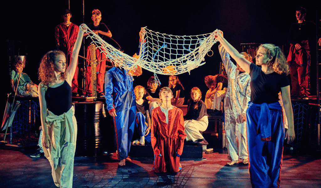 Background: Northcott stage. Black curtain backdrop. Set: Table staging. Oil barrels, nets and wind chimes. Foreground: Two performers wearing green (left) and blue (right) jumpsuits ted at their waists hold a net over another performer wearing a red jumpsuit. Behind them, a large group of performers, wearing black shirts and different coloured trousers, watch from different positions on the set.