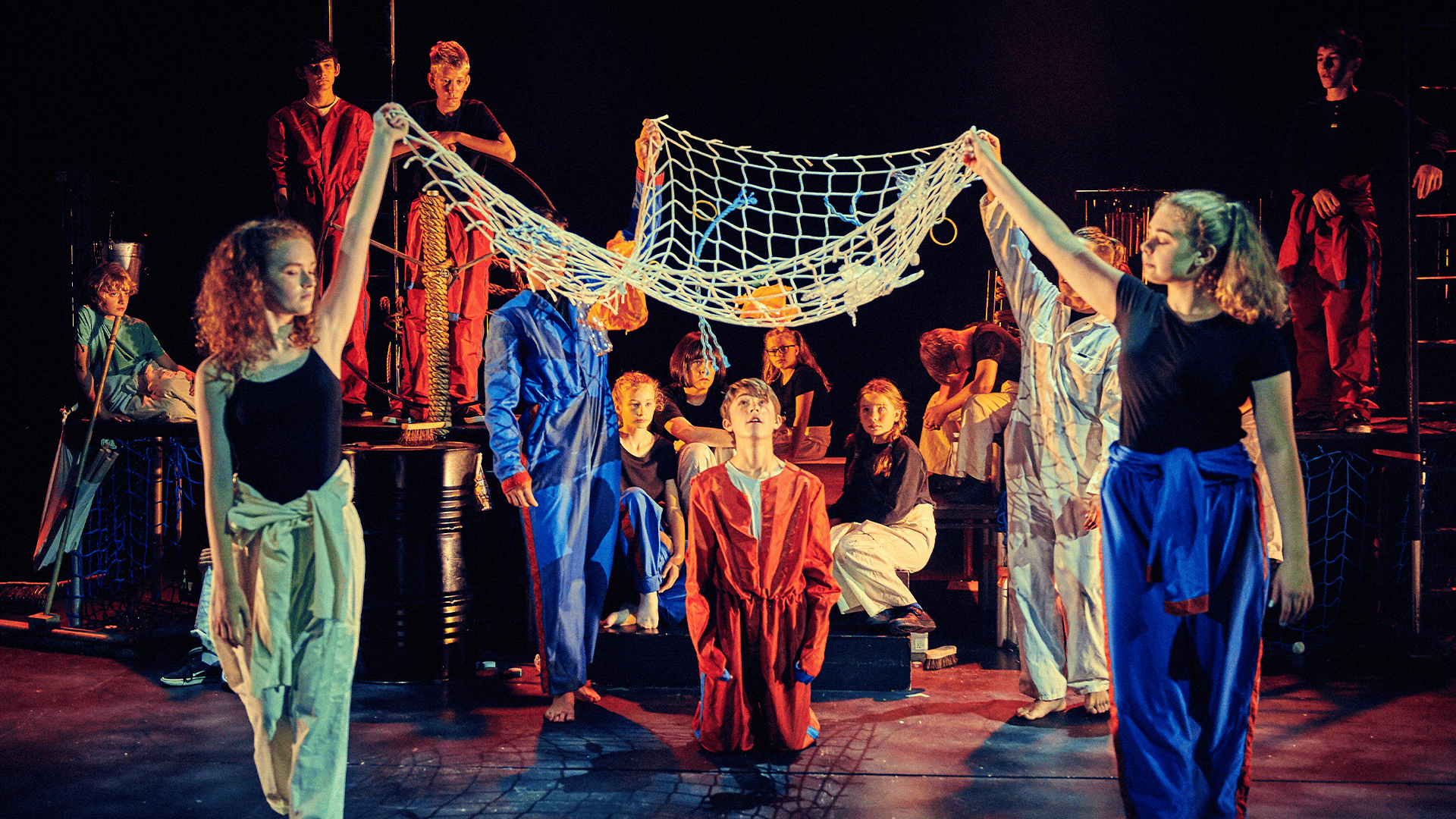 Background: Northcott stage. Black curtain backdrop. Set: Table staging. Oil barrels, nets and wind chimes. Foreground: Two performers wearing green (left) and blue (right) jumpsuits ted at their waists hold a net over another performer wearing a red jumpsuit. Behind them, a large group of performers, wearing black shirts and different coloured trousers, watch from different positions on the set.