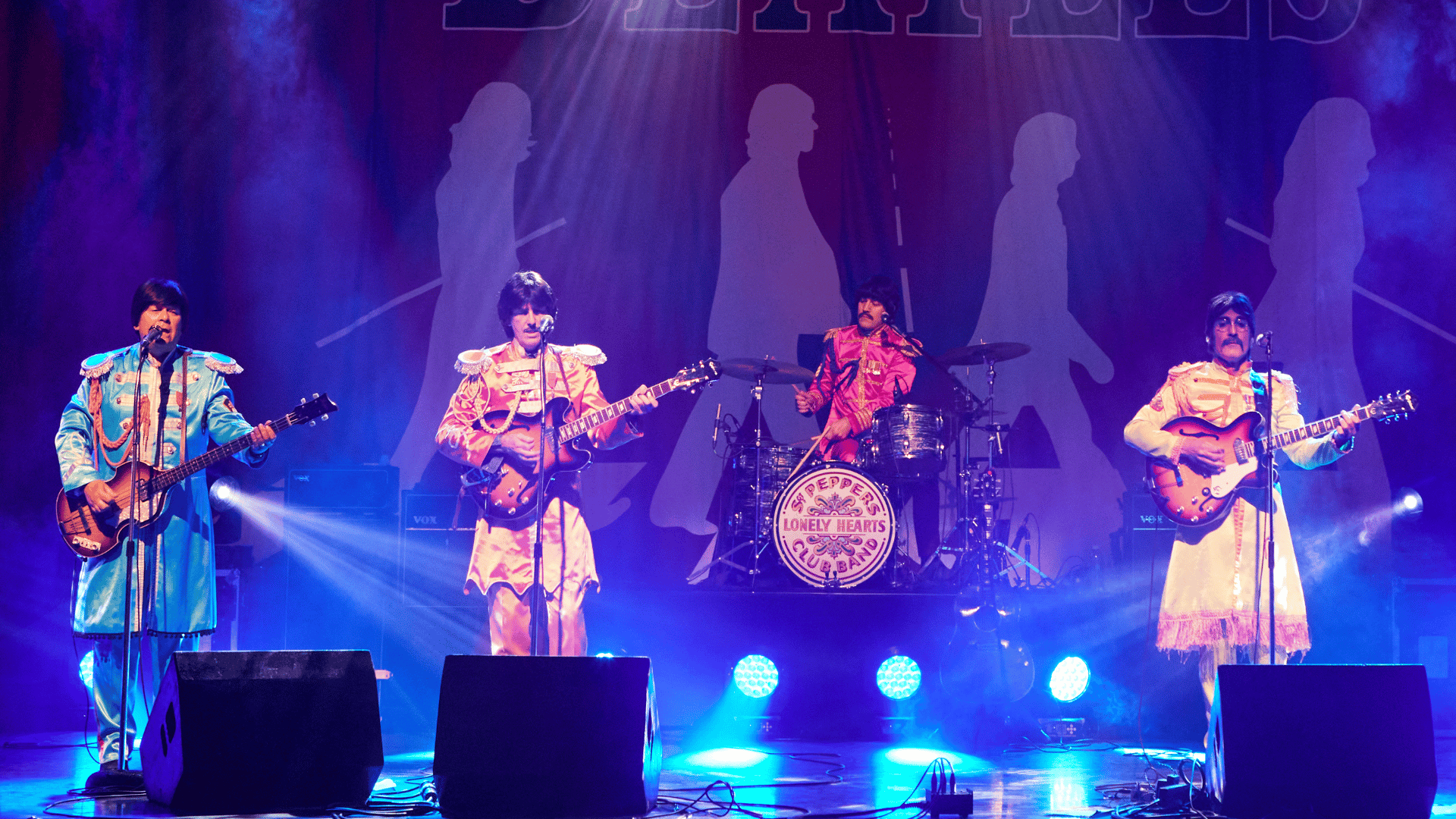 The Magic of the Beatles band stood in a line playing their instruments and singing at a live concert. They are wearing colourful outfits from the Sgt. Pepper album cover. From left to right: Paul, George, Ringo and John.